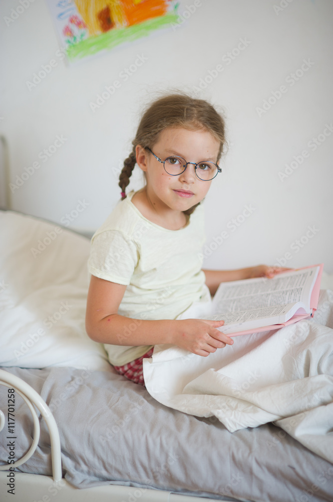 Little girl with glasses sitting in bed and holding an open book. Little girl with glasses sitting in bed and holding an open book.