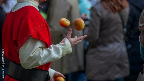 A young court jester is juggling with apples in among the visitors who are walking around and trying different foods. They came to see a battle of knights. Close-up shot.
 photo