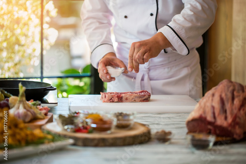 Hand holds bowl with salt. Tendeloin on cooking board. Spice needed for taste. Chef of restaurant prepares steak.