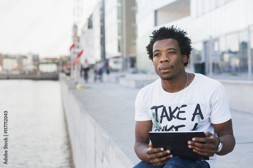 Thoughtful male college student holding digital tablet while sitting on retaining wall by river Thoughtful male college student holding digital tablet while sitting on retaining wall by river