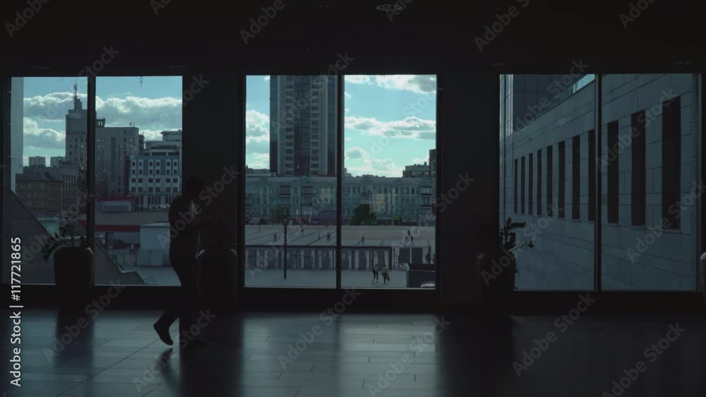Male athletic appearance is dark corridor with large windows looking into the phone. Office building. Outside seen as pedestrians walk. Male athletic appearance is dark corridor with large windows looking into the phone. Office building. Outside seen as pedestrians walk.