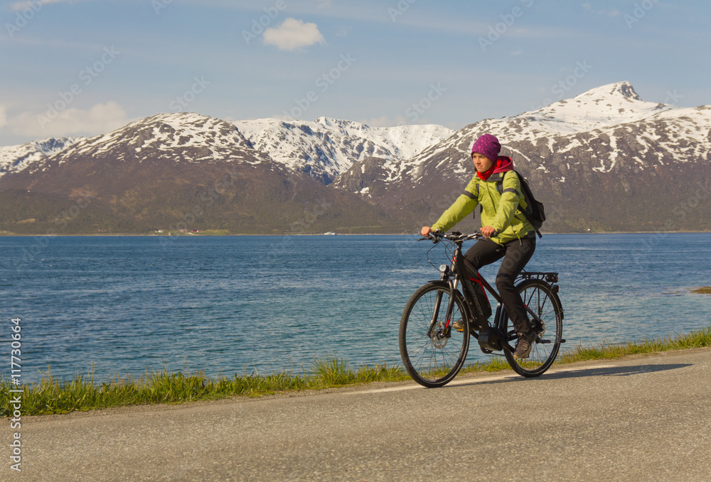Fototapeta premium junge Frau (Mädchen) fährt Fahrrad (E-Bike) auf Straße vor Bergen young woman (girl) riding a bicycle (e-bike) on road in front of mountains