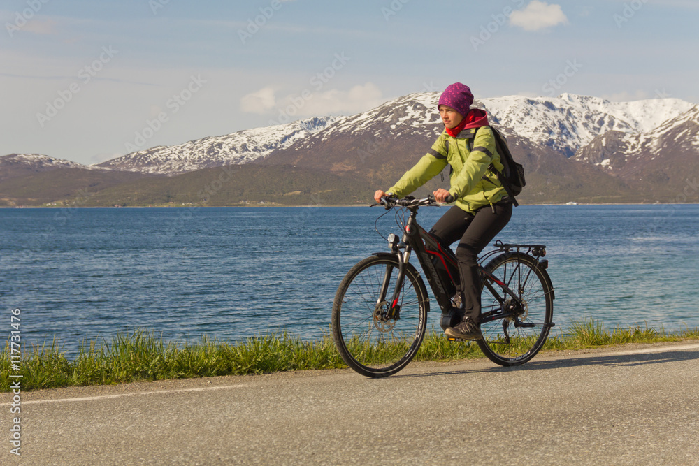 Obraz premium junge Frau (Mädchen) fährt Fahrrad (E-Bike) auf Straße vor Bergen young woman (girl) riding a bicycle (e-bike) on road in front of mountains 