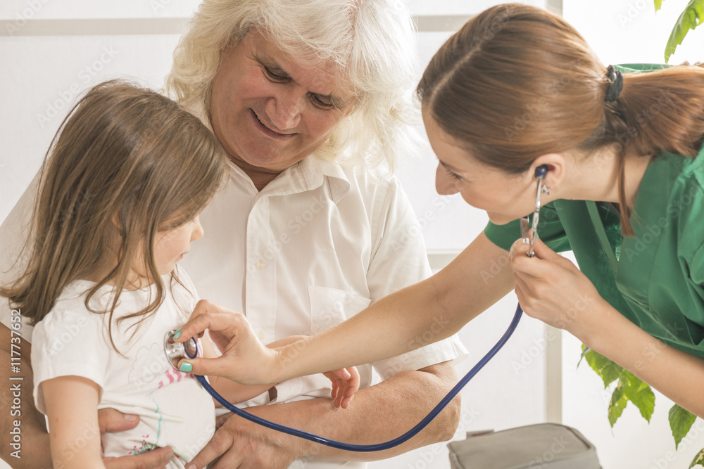 Grandfather holding his small granddaughter on his knees while she is being examined by female doctor in clinic Grandfather holding his small granddaughter on his knees while she is being examined by female doctor in clinic