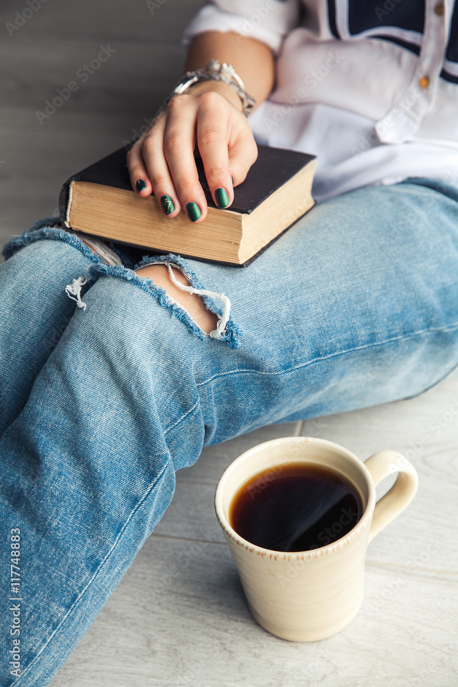 Obraz premium Young modern girl in torn jeans reading a book with a big cup of coffee. Fashion, lifestyle, lifestyle, recreation, education, hobbies.