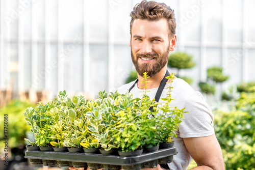 Portrait of a handsome gardener in black apron holding pots with plants in the greenhouse. Seller or worker in the plant shop
