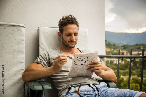 Young man sitting doing a crossword puzzle looking thoughtfully at a magazine, as he tries to think of the answer to the clue photo