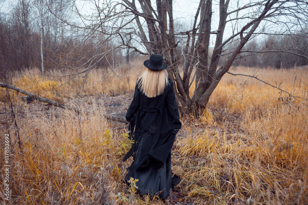 Portrait of young attractive woman in black coat and hat. She goes through the field. Autumn landscape, dry grass. Look back