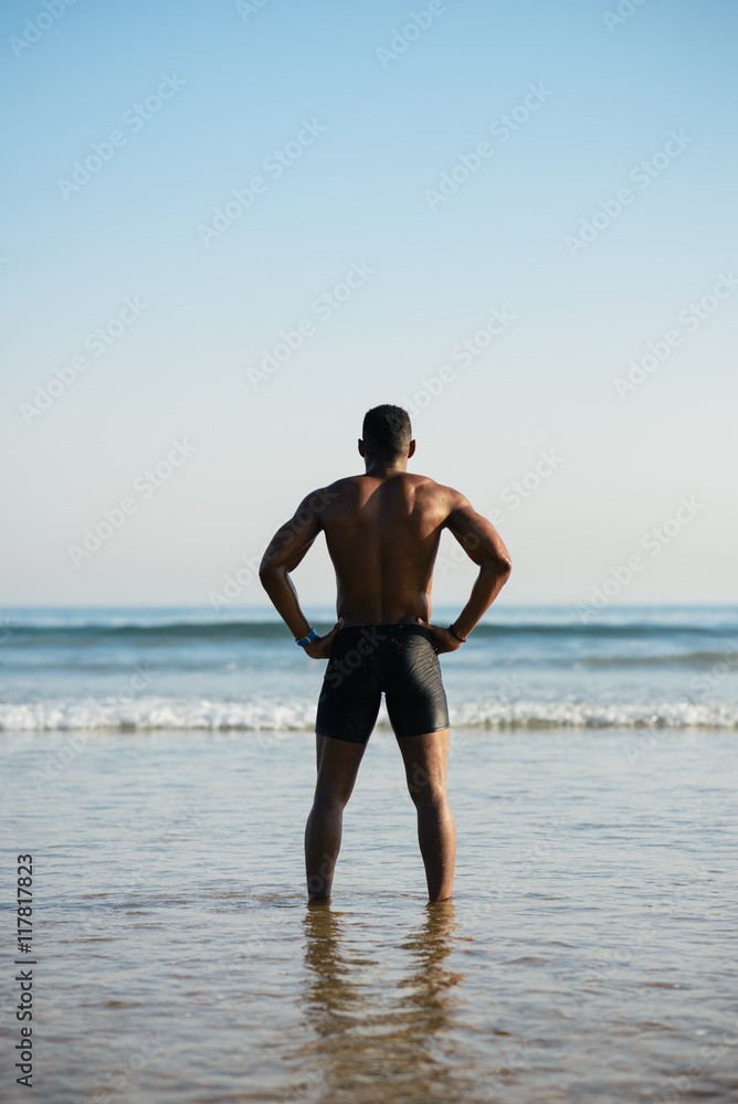 Back view of black fit sportsman looking the sea for motivation before swimming. Male swimmer ready for outdoor training into the ocean.