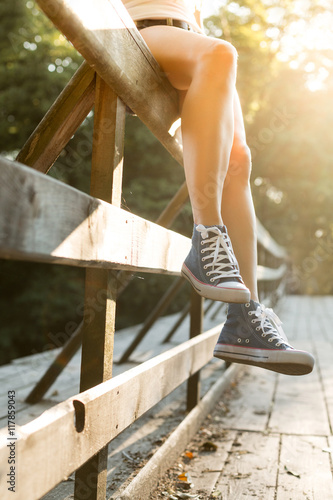 Young woman sitting on a bridge railing in jeans sneakers
