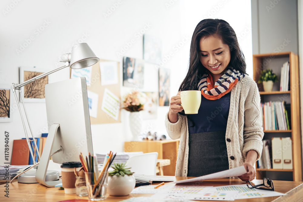 Smiling pretty female editor drinking coffee and reading document Smiling pretty female editor drinking coffee and reading document