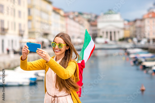 Young female traveler with backpack and italian flag making selfie photo with phone on Trieste city centre background. Traveling in Italy