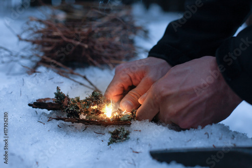 Man lighting a fire in a dark winter forest, preparing for an overnight sleep in nature, warming himself with DIY fire. Adventure, scouting, survival concept. . photo