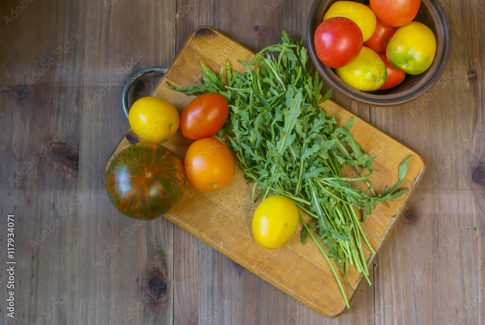 Fresh tomatoes and greens (arugula, parsley) on a cutting board on a wooden table. Near bowl with tomatoes Fresh tomatoes and greens (arugula, parsley) on a cutting board on a wooden table. Near bowl with tomatoes