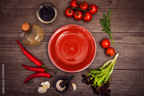 Fresh tomatoes, chili pepper and other spices and herbs around modern red plate in the center of wooden table and cloth napkin. Top view. Blank place for your text. Close-up.