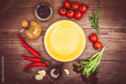 Fresh tomatoes, chili pepper and other spices and herbs around modern yellow plate in the center of wooden table. Top view. Copy space for your text. Close-up.