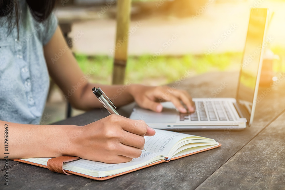 Fototapeta premium Female hands with pen writing notebook and using laptop on desk table at coffee shop.