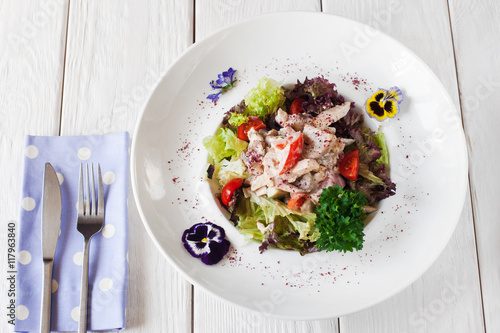 Chicken and mushroom salad on white plate, decorated with flowers. Restaurant meal of warm side dish with sauce, served with knife and fork on white wooden table
