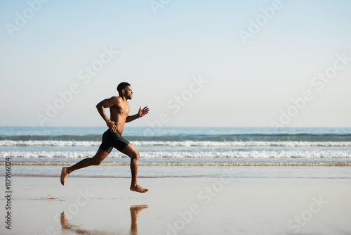 Black fit man running barefoot by the sea on the beach. Powerful runner training outdoor on summer.