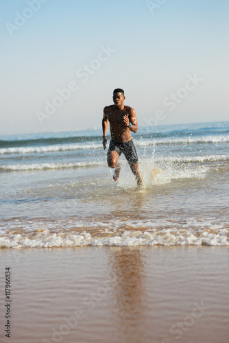 Man training at the beach. Black athlete running into the sea splashing. Runner sprinting at seashore during summer outdoor workout.