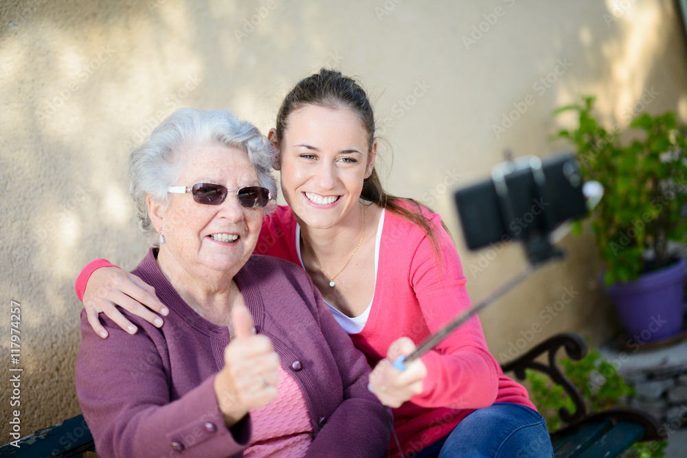 Fototapeta premium cheerful old woman in wheelchair making a selfie with her young granddaughter outdoor in hospital