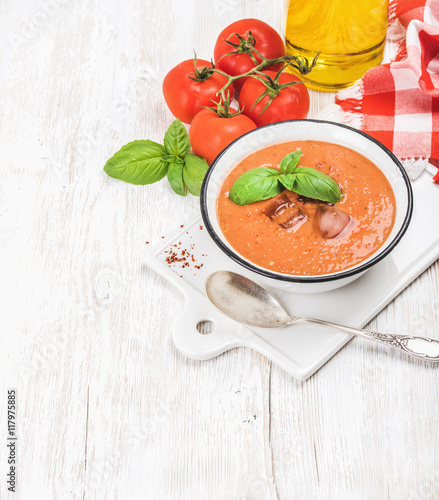Cold gazpacho soup in bowl with ice, hot pepper and basil served with fresh tomotoes on ceramic board over white wooden background, top view, selective focus, copy space, vertical composition