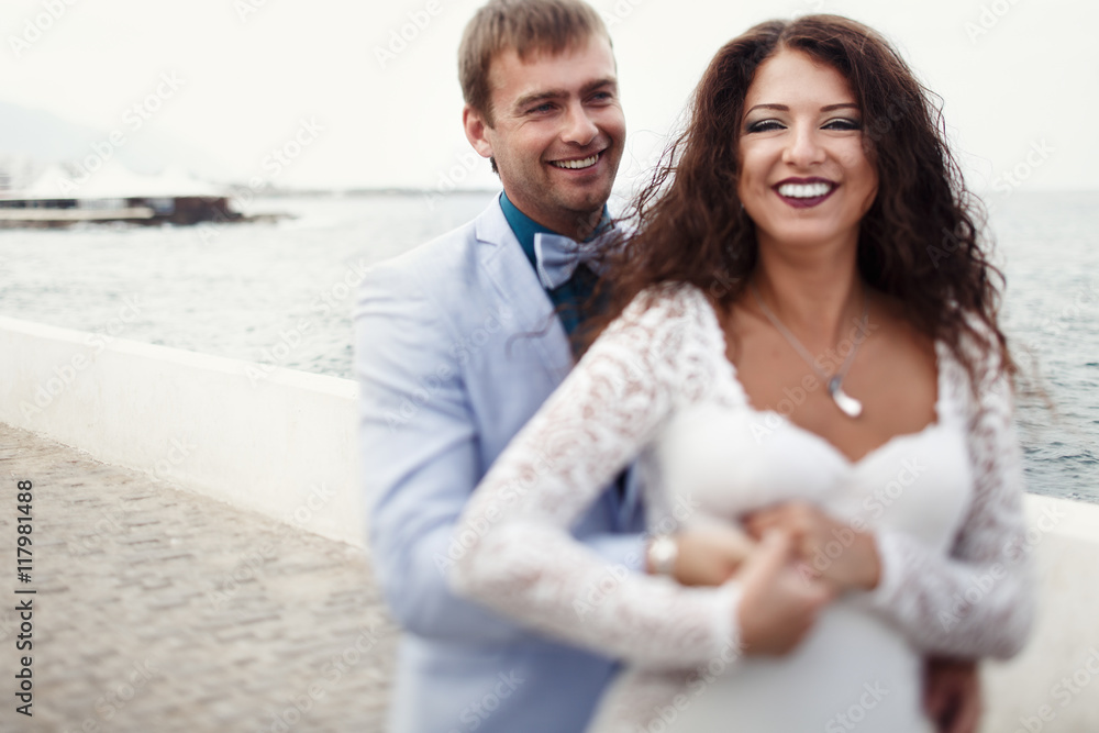 Obraz premium Groom holds bride's waist tightly while they pose by the sea