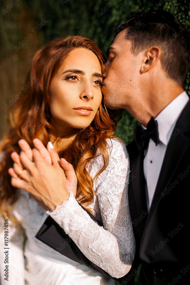Evening sun illuminates gorgeous bride's face while groom kisses Evening sun illuminates gorgeous bride's face while groom kisses