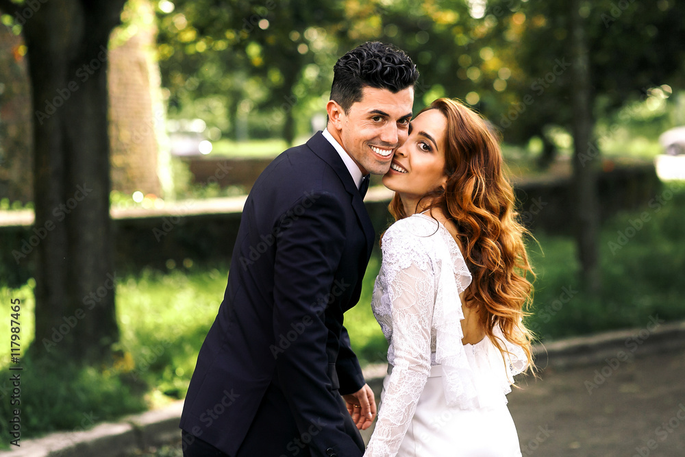 Curly bride leans to groom's cheek while posing in the park Curly bride leans to groom's cheek while posing in the park