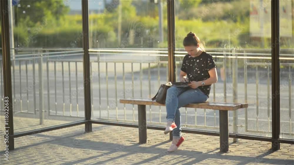 Young charming Caucasian female with trendy look use tablet while waiting transport on bus stop, woman sitting on a station against sunset background Young charming Caucasian female with trendy look use tablet while waiting transport on bus stop, woman sitting on a station against sunset background