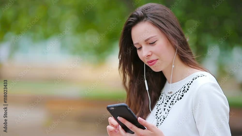 Young caucasian woman sending message by her smartphone outdoor in the park. Beautiful girl in sunglasses sitting on wooden bench using smartphone Young caucasian woman sending message by her smartphone outdoor in the park. Beautiful girl in sunglasses sitting on wooden bench using smartphone