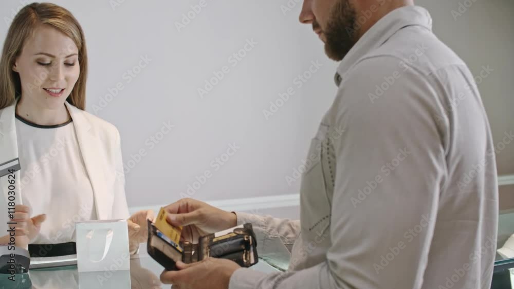 Man taking credit from his wallet and entering PIN on payment terminal while smiling shop assistant giving him branded paper bag with his purchase in jewelry store Man taking credit from his wallet and entering PIN on payment terminal while smiling shop assistant giving him branded paper bag with his purchase in jewelry store