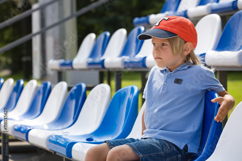 Obraz na plátně Portrait of cute blond kid boy sitting on sports tribune at the stadium and watching competition