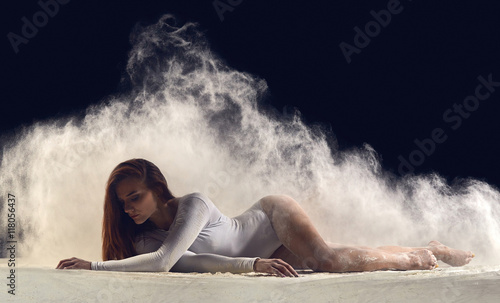 Fototapeta Naklejka Na Ścianę i Meble -  Dancer posing gracefully on the floor in a cloud of dust. Dance art and dancing in dust. Dancer in a white bathing suit.