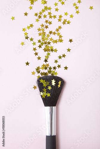 A cosmetics brush with gold sequin stars spilling out over a pastel pink background photo