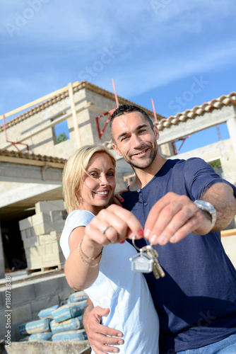 cheerful young happy couple holding home keys in front of contruction site of new house
