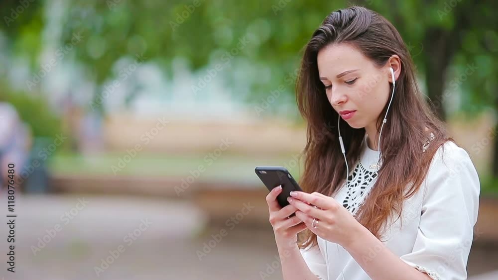 Young caucasian woman sending message by her smartphone outdoor in the park. Beautiful girl in sunglasses sitting on wooden bench using smartphone Young caucasian woman sending message by her smartphone outdoor in the park. Beautiful girl in sunglasses sitting on wooden bench using smartphone