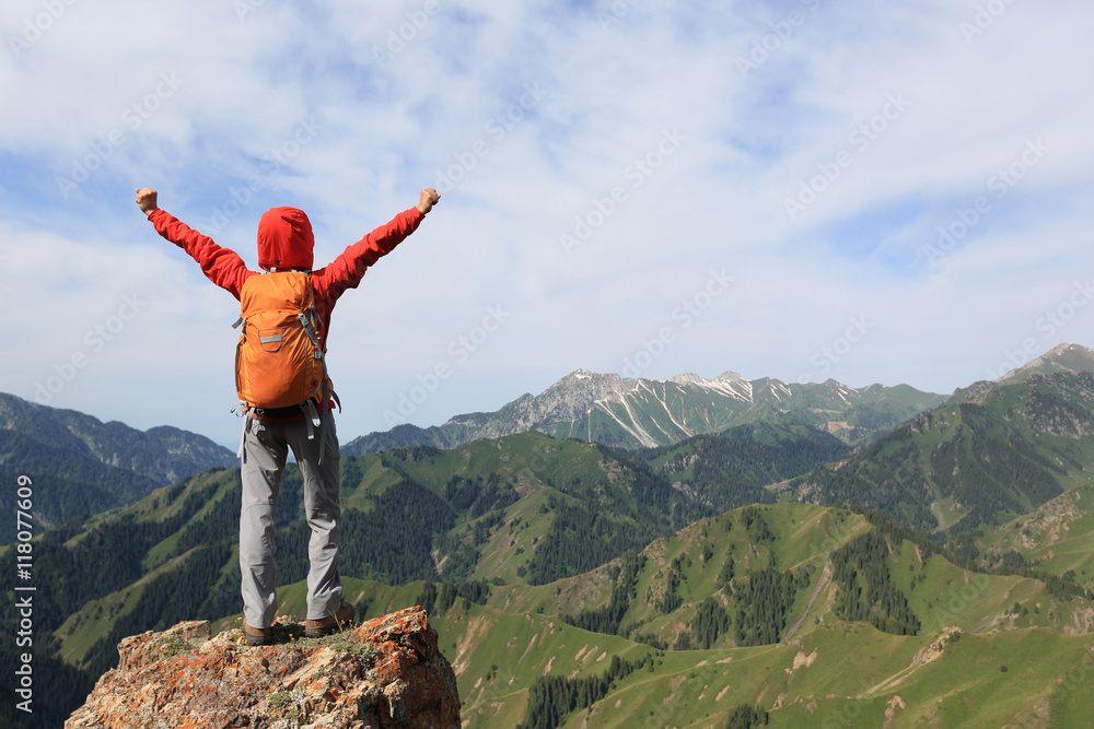 Obraz premium cheering young woman backpacker open arms on mountain peak cliff