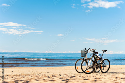 Two bicycles standing on the beach sand with blue cloudy sky bac