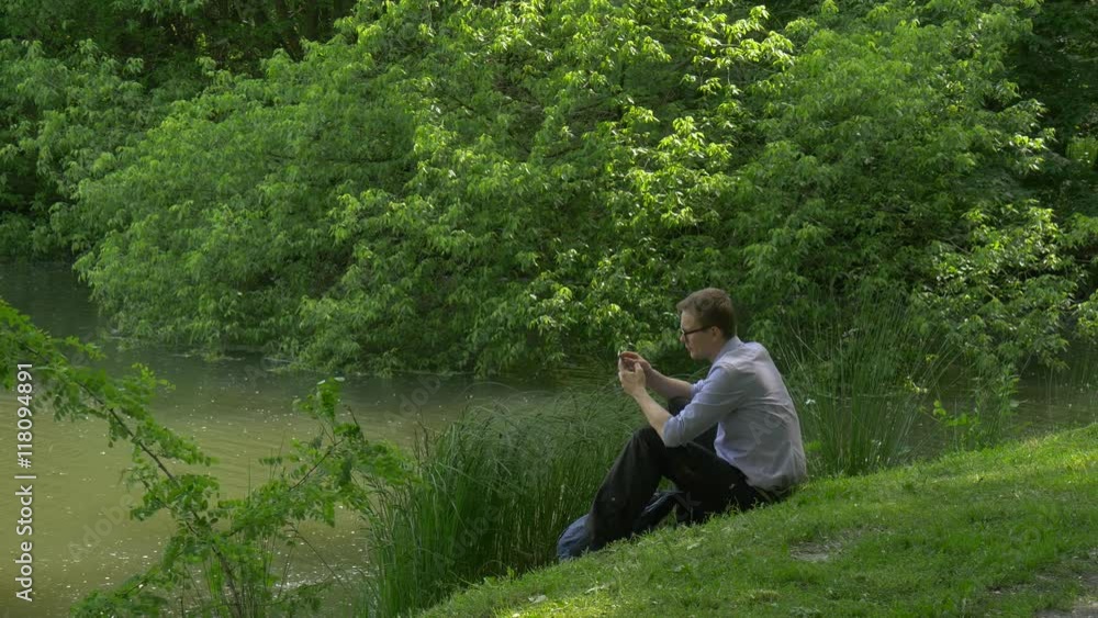 Businessman With Mobile Phone Sitting on Green Hill Holding a Smartphone Spends Time in Park Fresh Green Grass Trees Sunny Summer Day Businessman With Mobile Phone Sitting on Green Hill Holding a Smartphone Spends Time in Park Fresh Green Grass Trees Sunny Summer Day