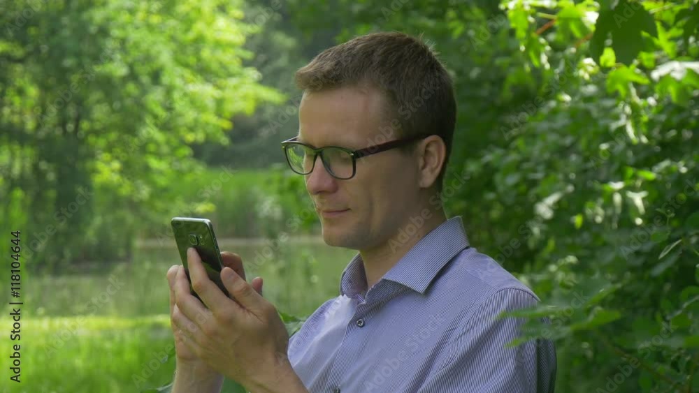 Man Clicks Mobile Phone in Park Smiling Texting Puts it Close to His Ear Sitting Among Green Trees Spend Time at the Nature Fresh Green Trees Sunny Summer Man Clicks Mobile Phone in Park Smiling Texting Puts it Close to His Ear Sitting Among Green Trees Spend Time at the Nature Fresh Green Trees Sunny Summer