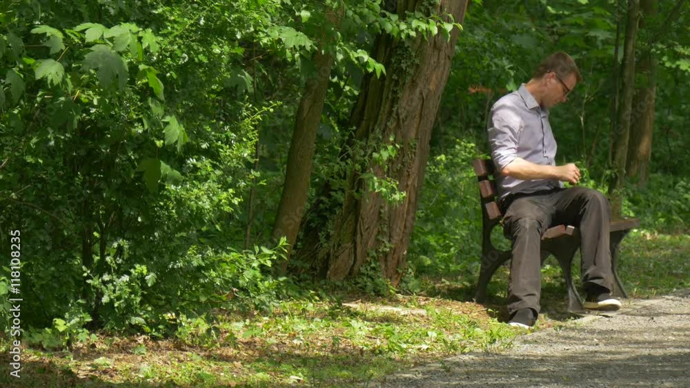 Man Takes Mobile Phone Gets up of Bench in Park Walking Away Holding a Smartphone Man in Glasses Spend Time at the Nature Fresh Green Trees Sunny Day Man Takes Mobile Phone Gets up of Bench in Park Walking Away Holding a Smartphone Man in Glasses Spend Time at the Nature Fresh Green Trees Sunny Day