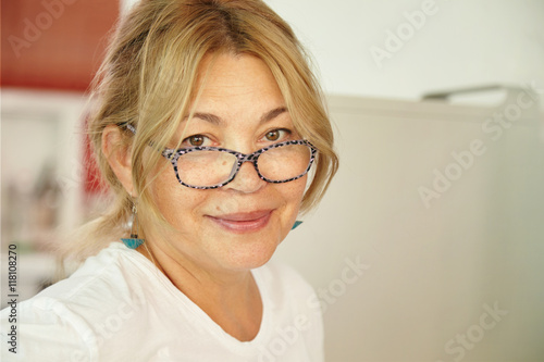Good-looking Caucasian middle-aged female with blond hair and kind eyes, looking with tender and friendly smile while waiting for her grandchildren to visit her at home during summer vacations