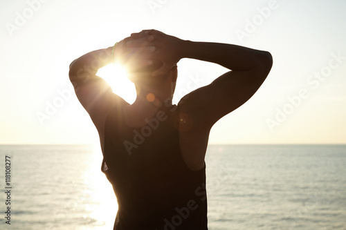 Silhouette of young African man wearing sleeveless top clasping hands behind his head, admiring beautiful view of sun setting over ocean, listening to sounds of sea on summer evening. Flare sun