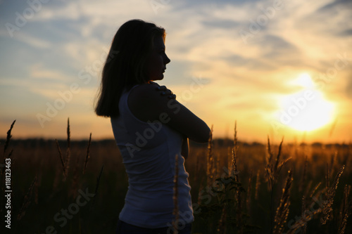 Silhouette young woman hugging herself in a wheat field. He looks into the distance. Sunset.