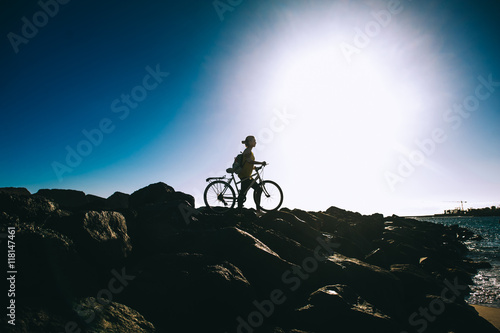 Young woman with backpack standing on the shore near his bike an