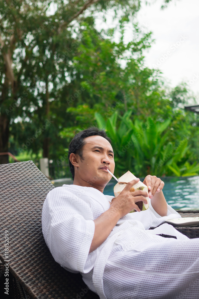 Fototapeta premium man drinking a coconut juice on deck chairs next to a swimming pool 