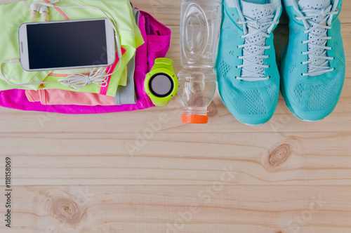 Top view of outfit for runner on wooden background: bottle of water, gps watch, running shoes, running waist bag, shorts, shirt, and sport bra. Horizontal orientation.