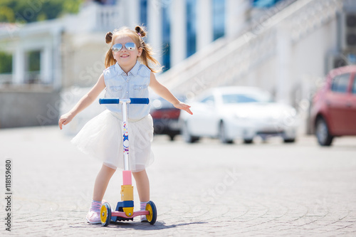 Little girl with blond hair,hair in two ponytails,in mirrored sun glasses,a light blue sleeveless shirt and white skirt, the one riding on the scooter in the city in the fresh air in summer