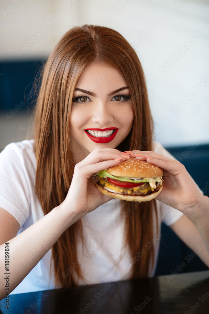 Beautiful woman with red long straight hair,bright make-up,brown eyes,red lipstick,long eyelashes,pink nail polish in the hands holding a large hamburger, sitting at a table in a cafe,dinner alone Beautiful woman with red long straight hair,bright make-up,brown eyes,red lipstick,long eyelashes,pink nail polish in the hands holding a large hamburger, sitting at a table in a cafe,dinner alone